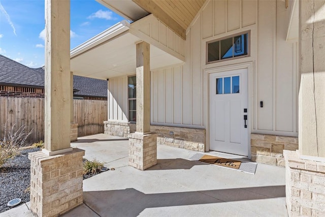 view of exterior entry with stone siding, fence, board and batten siding, and a patio