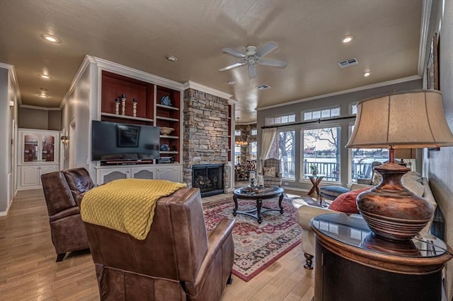 living area with visible vents, light wood-style flooring, ceiling fan, crown molding, and a stone fireplace