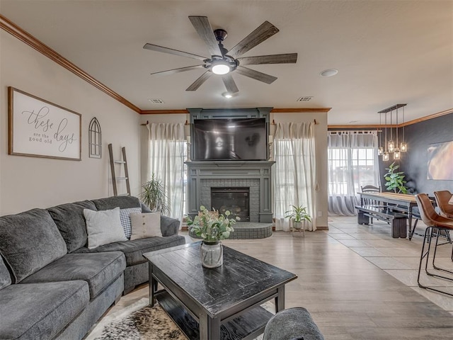 living room with visible vents, ornamental molding, light wood-style floors, a brick fireplace, and a ceiling fan