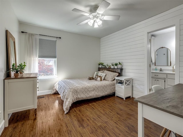 bedroom with wooden walls, dark wood finished floors, a ceiling fan, ensuite bathroom, and a sink