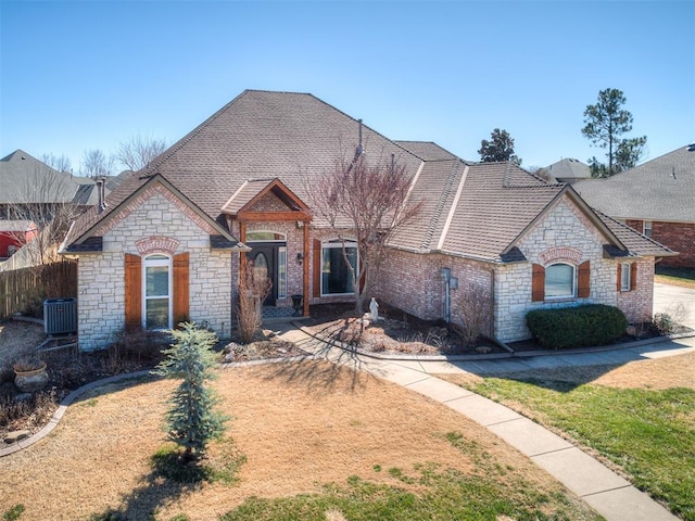 french country inspired facade featuring a front lawn, roof with shingles, fence, and central air condition unit