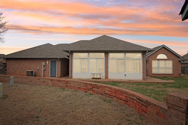 back of property at dusk featuring brick siding, a shingled roof, fence, a sunroom, and a yard