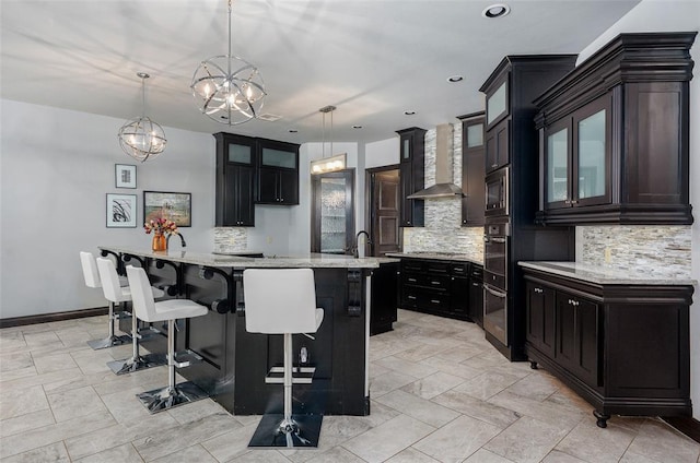 kitchen with a breakfast bar, a center island with sink, tasteful backsplash, glass insert cabinets, and wall chimney exhaust hood