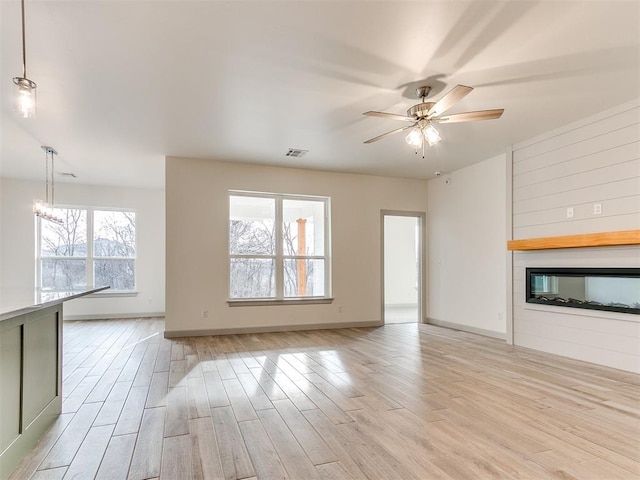 unfurnished living room with light wood finished floors, visible vents, a glass covered fireplace, baseboards, and ceiling fan with notable chandelier