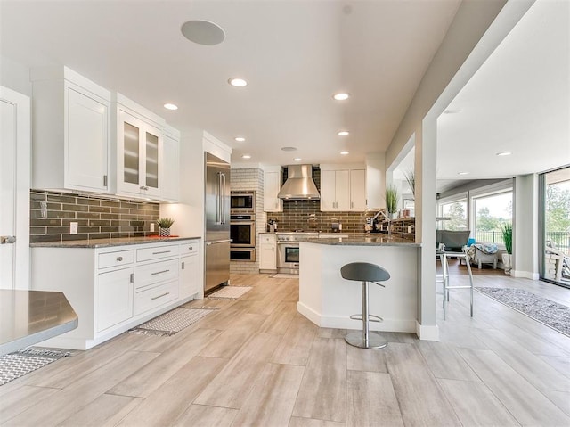 kitchen featuring a kitchen bar, white cabinets, wall chimney exhaust hood, and built in appliances