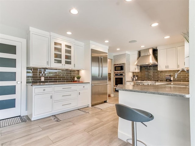 kitchen featuring white cabinets, wall chimney exhaust hood, glass insert cabinets, built in appliances, and a kitchen bar