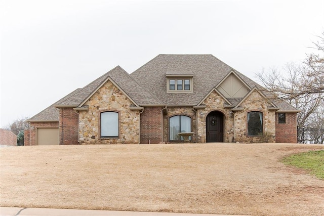 french country inspired facade featuring a garage, driveway, a shingled roof, and brick siding