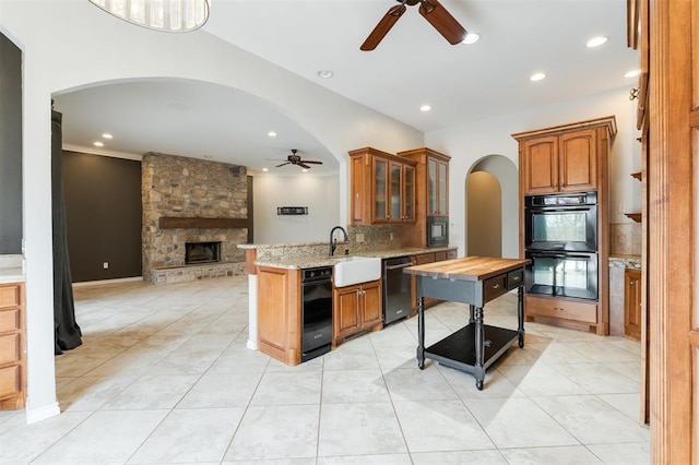 kitchen with arched walkways, brown cabinetry, a sink, a stone fireplace, and black appliances