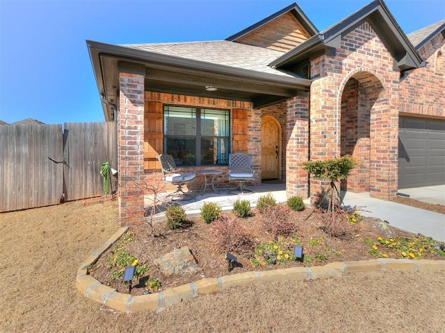 view of front of house with a porch, an attached garage, brick siding, fence, and roof with shingles