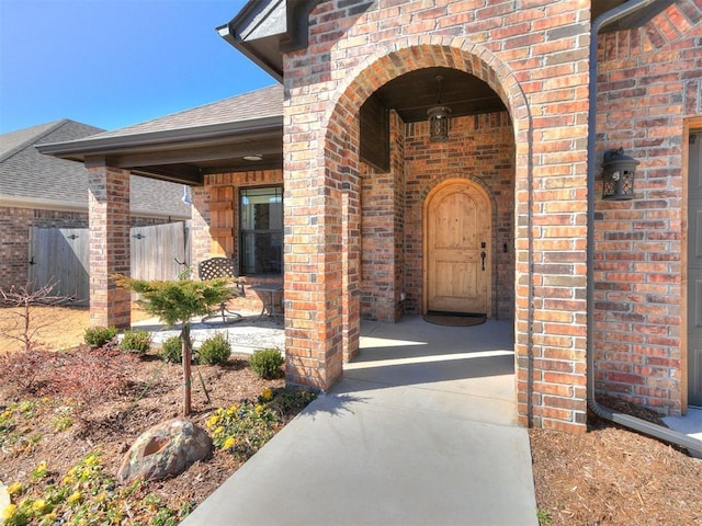 doorway to property featuring a shingled roof, fence, and brick siding