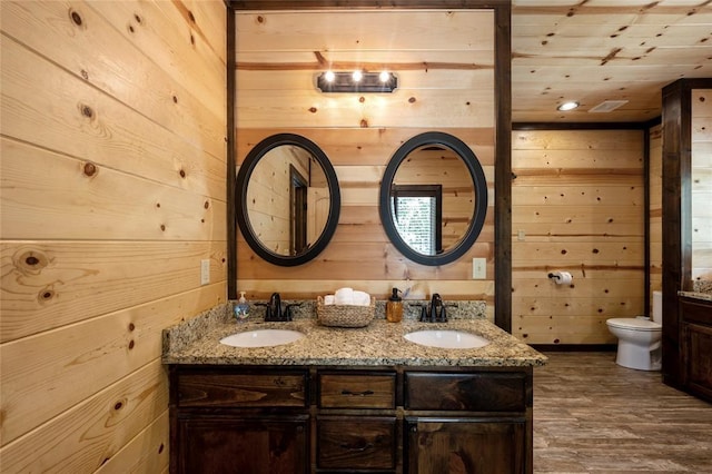 bathroom with double vanity, wooden walls, toilet, and a sink