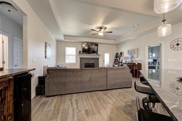 living area featuring a stone fireplace, a tray ceiling, a ceiling fan, and light wood-style floors