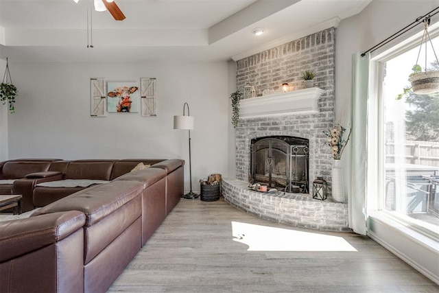 living room with a ceiling fan, a tray ceiling, a brick fireplace, and wood finished floors