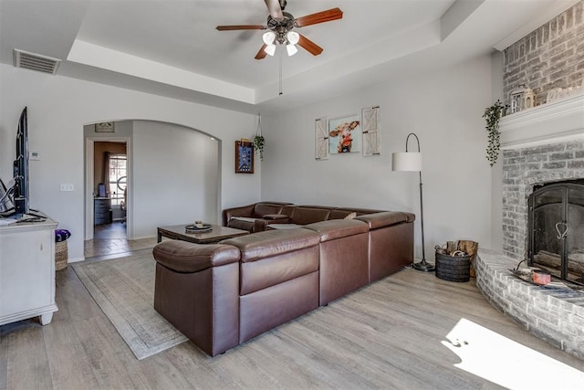 living room featuring a tray ceiling, arched walkways, visible vents, light wood-style flooring, and a brick fireplace