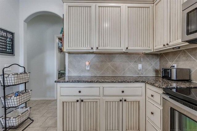 kitchen featuring arched walkways, light tile patterned flooring, stainless steel appliances, dark stone counters, and tasteful backsplash