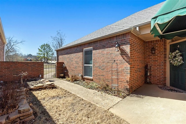 view of side of property with a gate, brick siding, fence, and roof with shingles