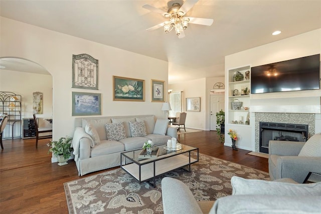 living room featuring arched walkways, a fireplace with flush hearth, wood finished floors, a ceiling fan, and built in features