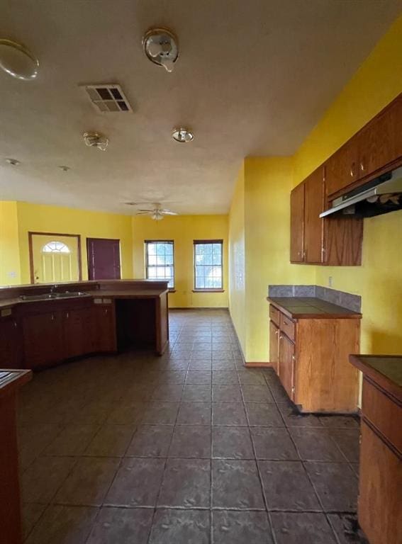 kitchen with visible vents, brown cabinetry, a ceiling fan, under cabinet range hood, and dark tile patterned floors