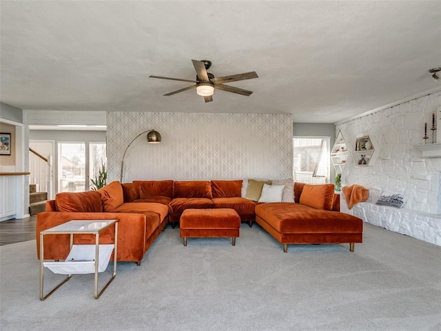 carpeted living room with stairs, a stone fireplace, a ceiling fan, and wallpapered walls