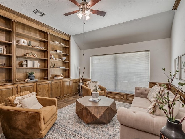 living area featuring a textured ceiling, wood finished floors, visible vents, vaulted ceiling, and wainscoting