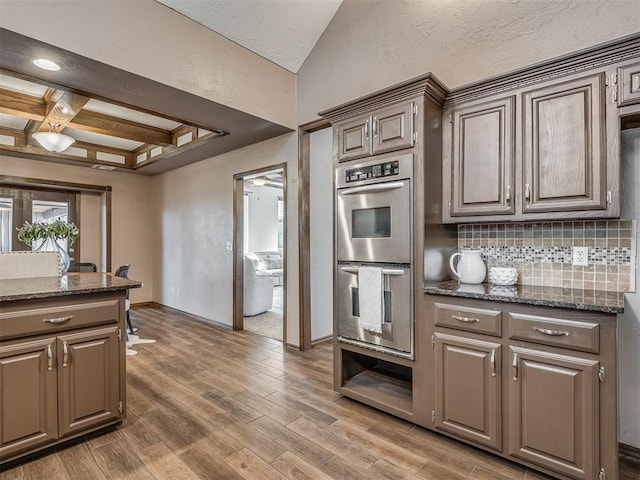 kitchen featuring light wood finished floors, tasteful backsplash, coffered ceiling, dark stone countertops, and stainless steel double oven