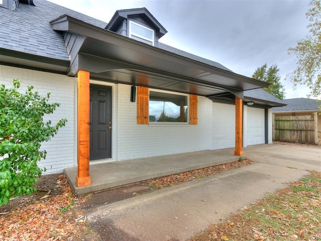 doorway to property featuring a shingled roof, concrete driveway, an attached garage, fence, and brick siding