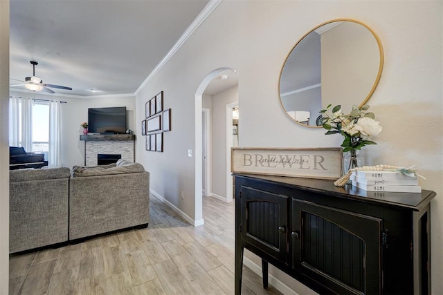 living area featuring arched walkways, a stone fireplace, baseboards, ornamental molding, and light wood-type flooring