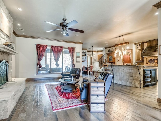 living room with visible vents, hardwood / wood-style flooring, ceiling fan, crown molding, and a stone fireplace