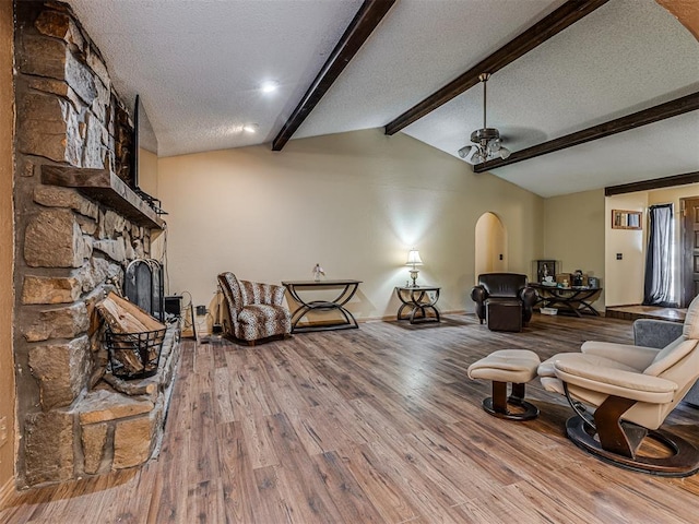 living area featuring lofted ceiling with beams, a stone fireplace, wood finished floors, arched walkways, and a textured ceiling