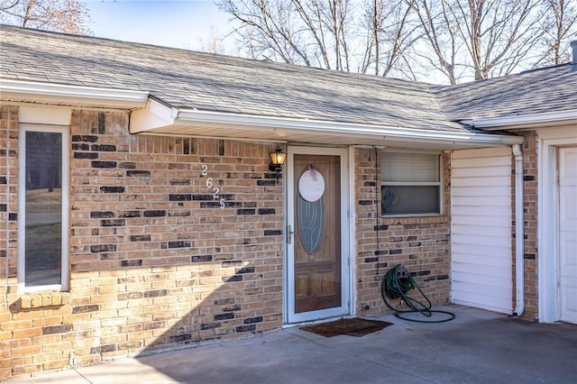 doorway to property featuring a garage, brick siding, and roof with shingles