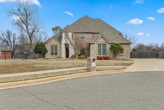 french country inspired facade featuring brick siding, fence, roof with shingles, stone siding, and driveway