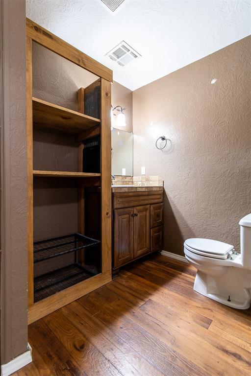 half bath with vanity, visible vents, hardwood / wood-style flooring, toilet, and a textured wall