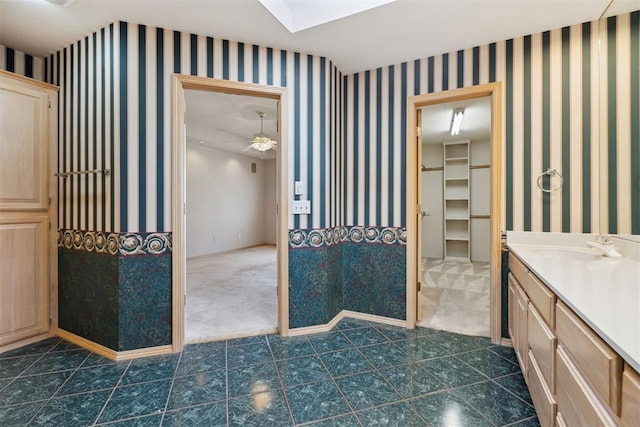 full bathroom with vanity, a ceiling fan, baseboards, wallpapered walls, and a walk in closet