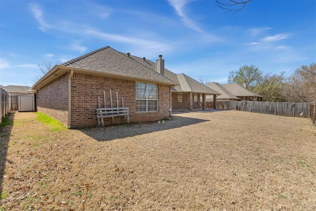 back of property with an outbuilding, a fenced backyard, roof with shingles, brick siding, and a chimney