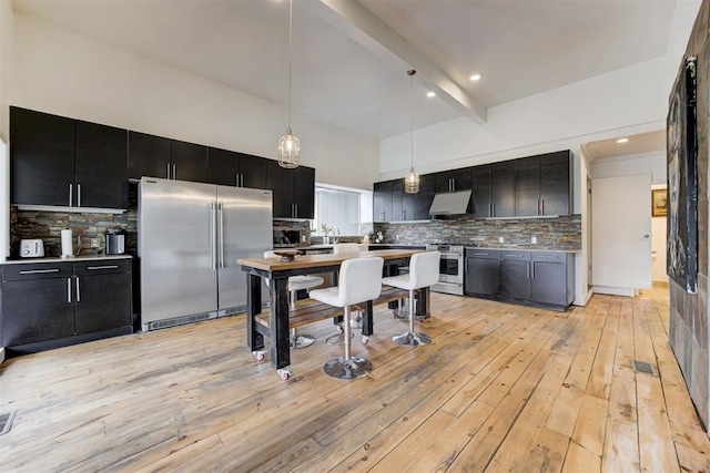 kitchen featuring beamed ceiling, under cabinet range hood, dark cabinetry, appliances with stainless steel finishes, and light wood finished floors