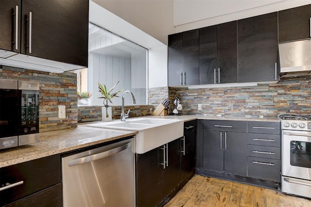 kitchen with a sink, backsplash, stainless steel appliances, light wood finished floors, and light stone countertops