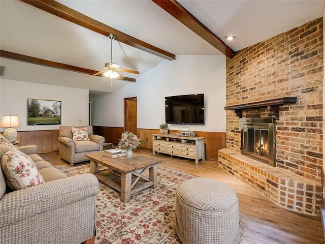 living room featuring a wainscoted wall, a ceiling fan, lofted ceiling with beams, wood finished floors, and a brick fireplace