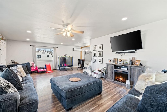 living area with a wall unit AC, a ceiling fan, wood finished floors, recessed lighting, and a warm lit fireplace