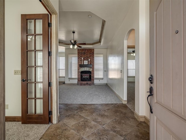 foyer with a brick fireplace, ceiling fan, a tray ceiling, arched walkways, and dark colored carpet