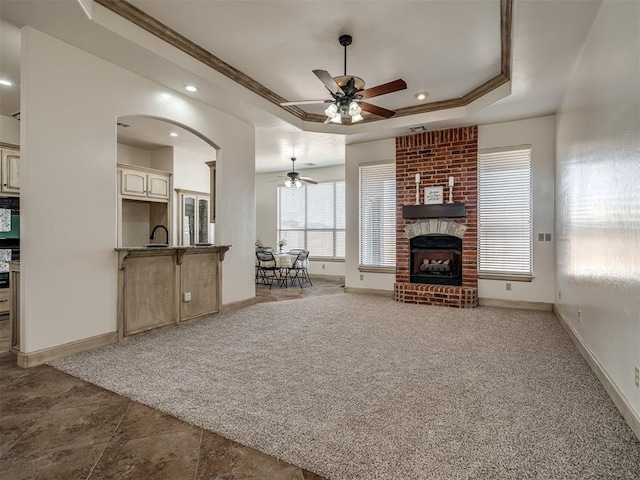 unfurnished living room featuring a brick fireplace, baseboards, light colored carpet, ornamental molding, and a raised ceiling