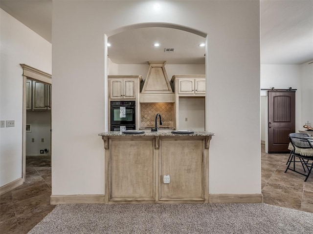 kitchen with dobule oven black, light stone counters, a sink, a barn door, and custom exhaust hood