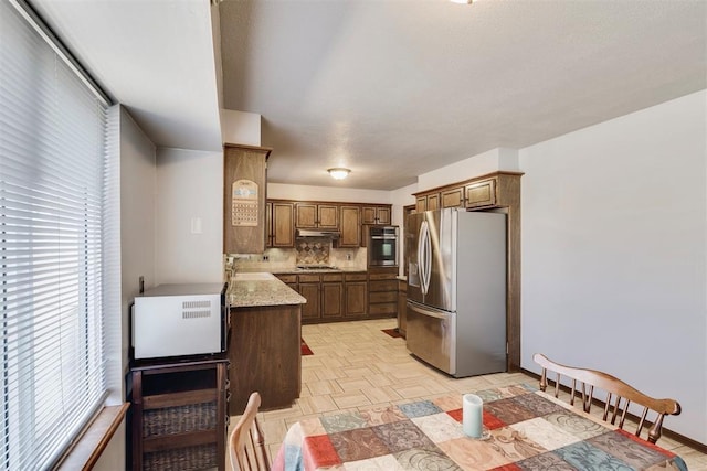 kitchen featuring brown cabinets, a sink, under cabinet range hood, backsplash, and appliances with stainless steel finishes