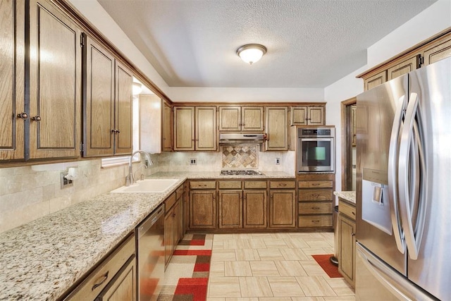 kitchen with brown cabinets, under cabinet range hood, a sink, tasteful backsplash, and stainless steel appliances