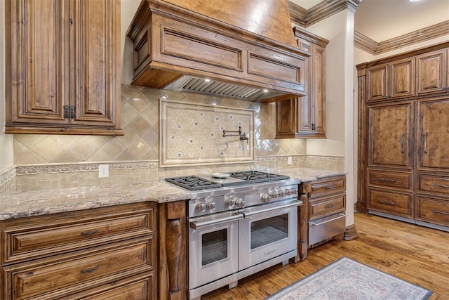 kitchen with light stone countertops, premium range hood, light wood-type flooring, ornamental molding, and range with two ovens