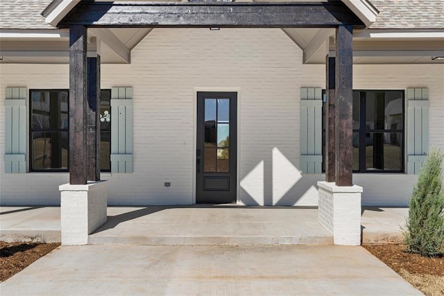 doorway to property featuring brick siding, a porch, and roof with shingles