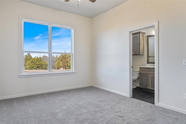 unfurnished bedroom featuring dark colored carpet, baseboards, ensuite bath, and a ceiling fan