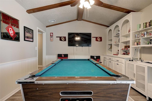 rec room featuring a ceiling fan, a wainscoted wall, vaulted ceiling with beams, light wood-style floors, and a textured ceiling