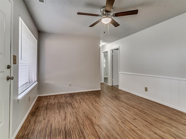 empty room featuring a ceiling fan, a textured ceiling, wood finished floors, wainscoting, and baseboards