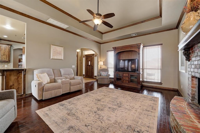 living area featuring a ceiling fan, arched walkways, dark wood-type flooring, crown molding, and a brick fireplace