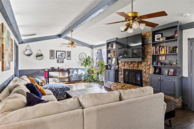 living area featuring built in features, a ceiling fan, vaulted ceiling with beams, a stone fireplace, and dark wood-type flooring